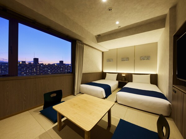 A hotel room with two beds, a small table, and a view of a cityscape at dusk. - Village Kyoto close by Shijo Omiya station (Kyoto)