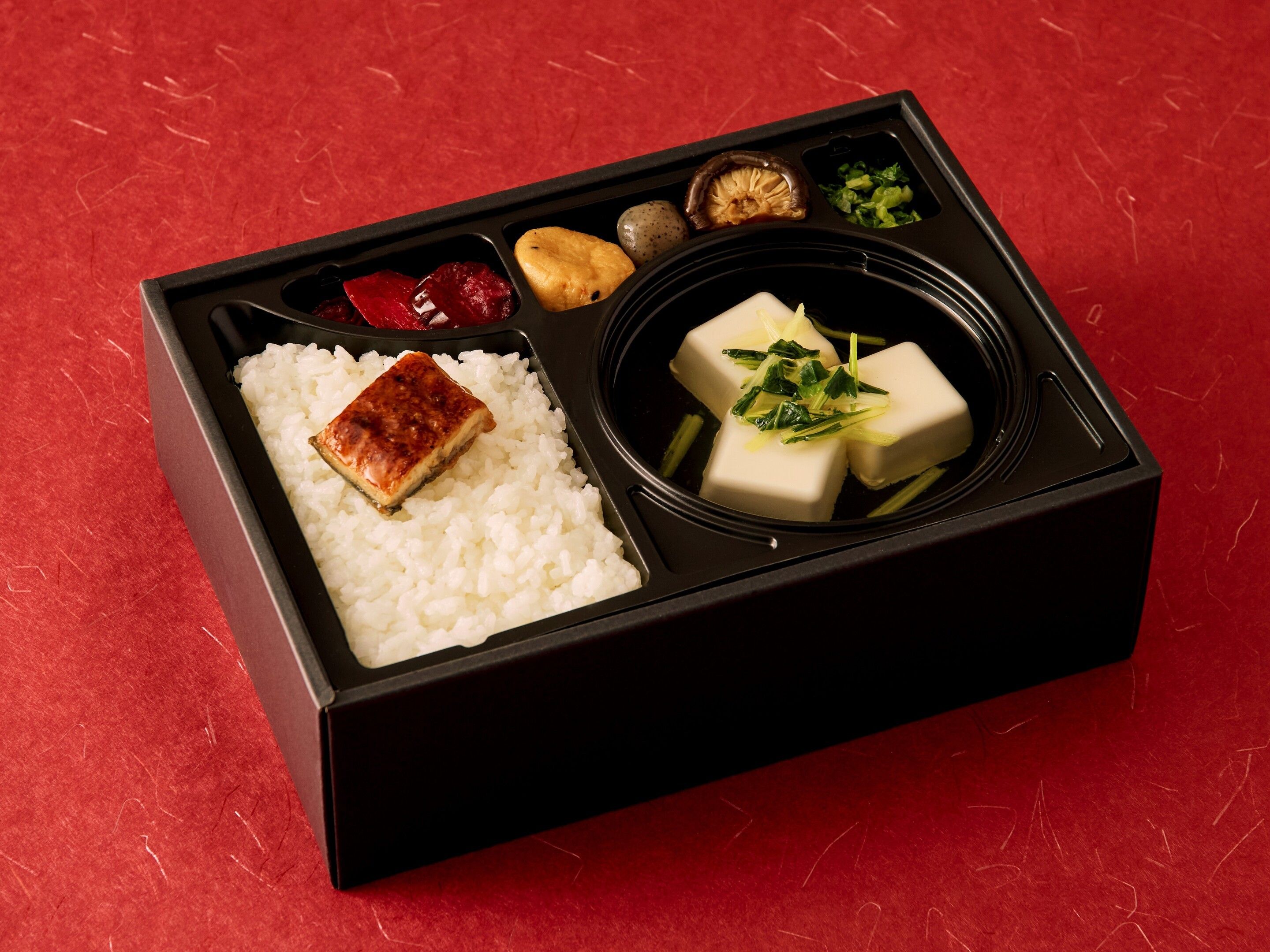 Une boîte à bento contenant du riz, du tofu, des légumes marinés et une petite portion de viande, le tout sur un fond rouge.