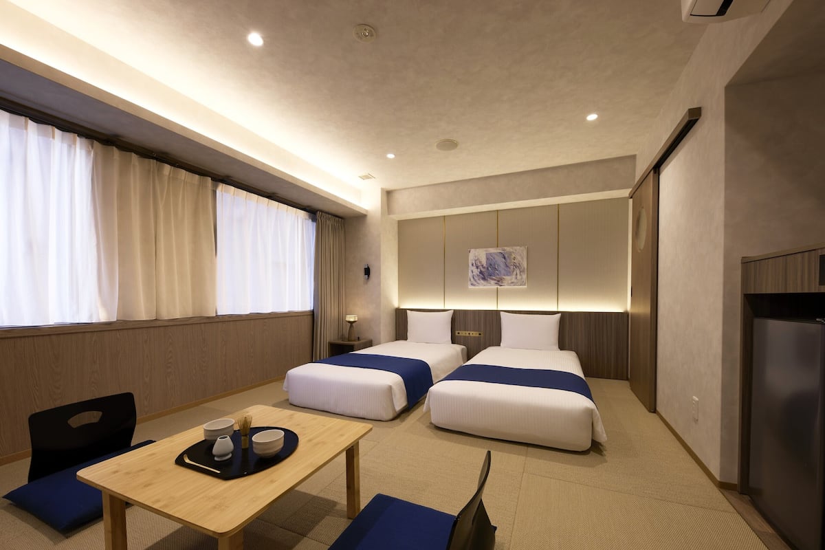 A hotel room with two beds, a dining table, a small kitchen area, and a window with curtains.