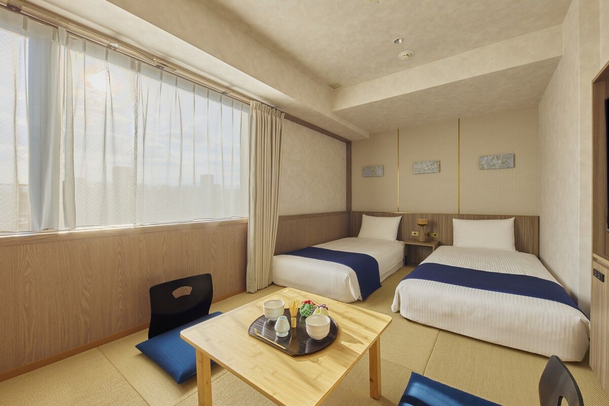 A hotel room with two beds, a TV, a small table with a tray of tea, and a window with curtains.
