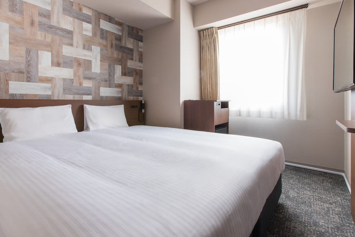 Een hotelkamer met een groot bed, een houten hoofdbord, een bureau met een stoel, een telefoon en een raam met gordijnen.