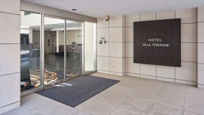 Eine moderne Hotelhalle mit Glastür, einer dunkel gefliesten Wand mit der Aufschrift „HOTEL VILLA FONTAINE“ und einem Fliesenboden.