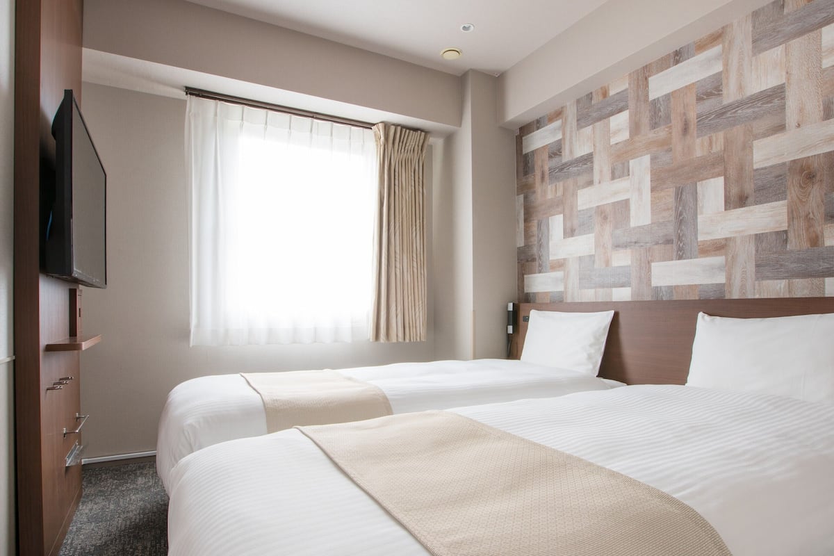 Een hotelkamer met twee bedden, een aan de muur bevestigde televisie, een raam met gordijnen en een houten wandpaneel.