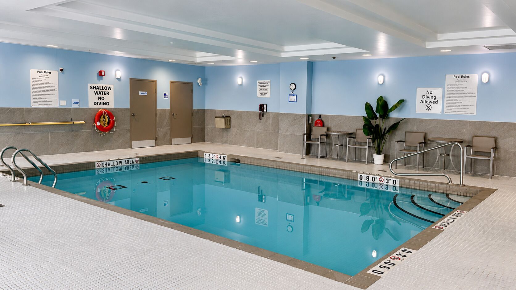 Une piscine intérieure avec des panneaux indiquant clairement les règles à respecter, des panneaux d’avertissement interdisant la plongée dans les zones peu profondes, et un poste de surveillance pour le sauveteur.