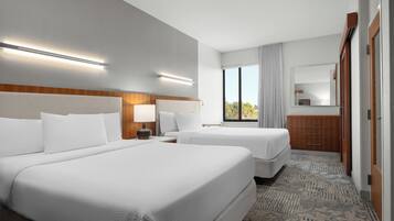 A hotel room with two beds, a wooden headboard, a nightstand with a lamp, a dresser, and a large window with curtains.