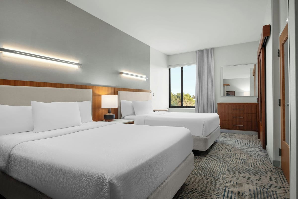 A hotel room with two beds, a wooden headboard, a nightstand with a lamp, a dresser, and a large window with a view of trees.