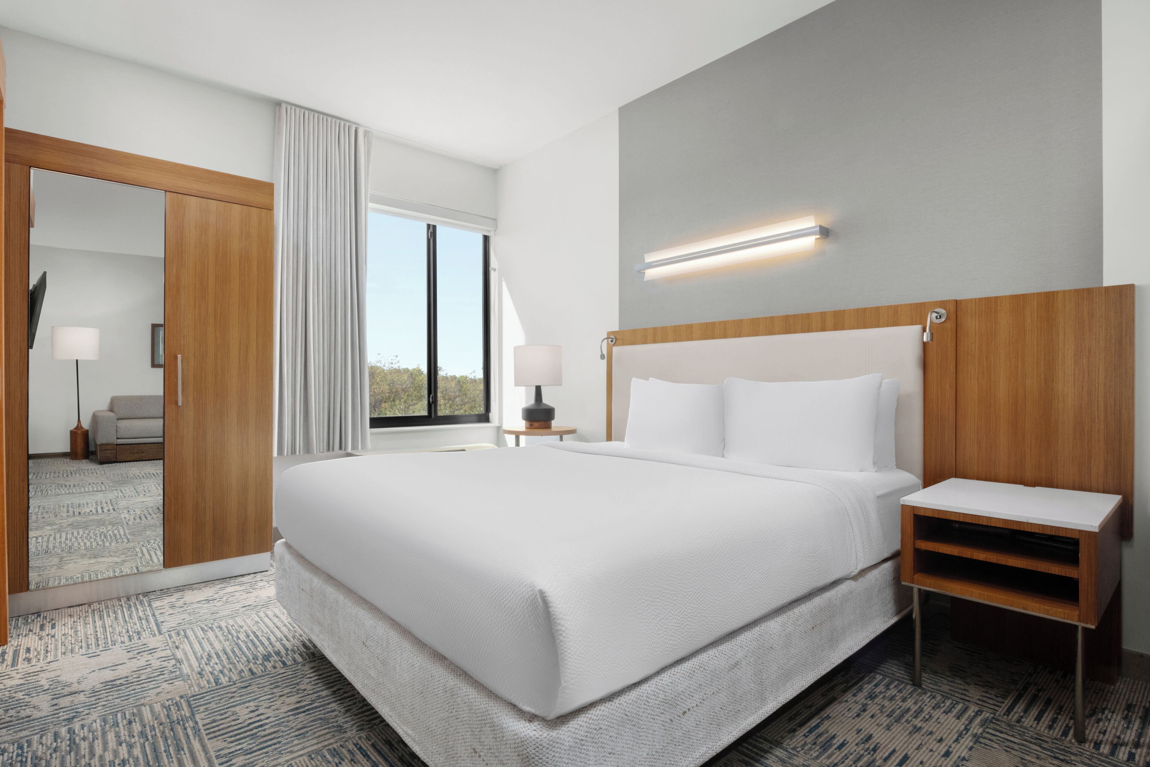 A hotel room with a large bed, a bedside table, a lamp, a mirror, and a view of the outdoors.