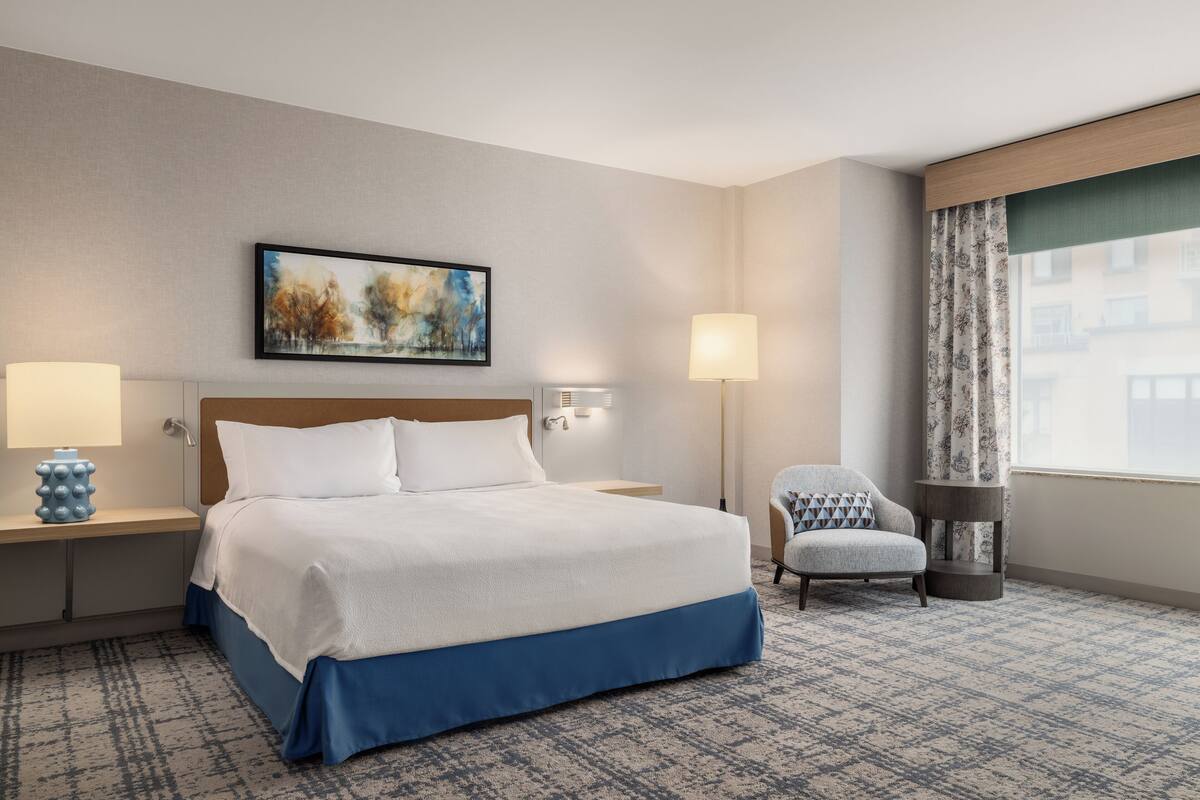 A hotel room with a large bed, two bedside lamps, a chair, a painting, and a window with curtains.