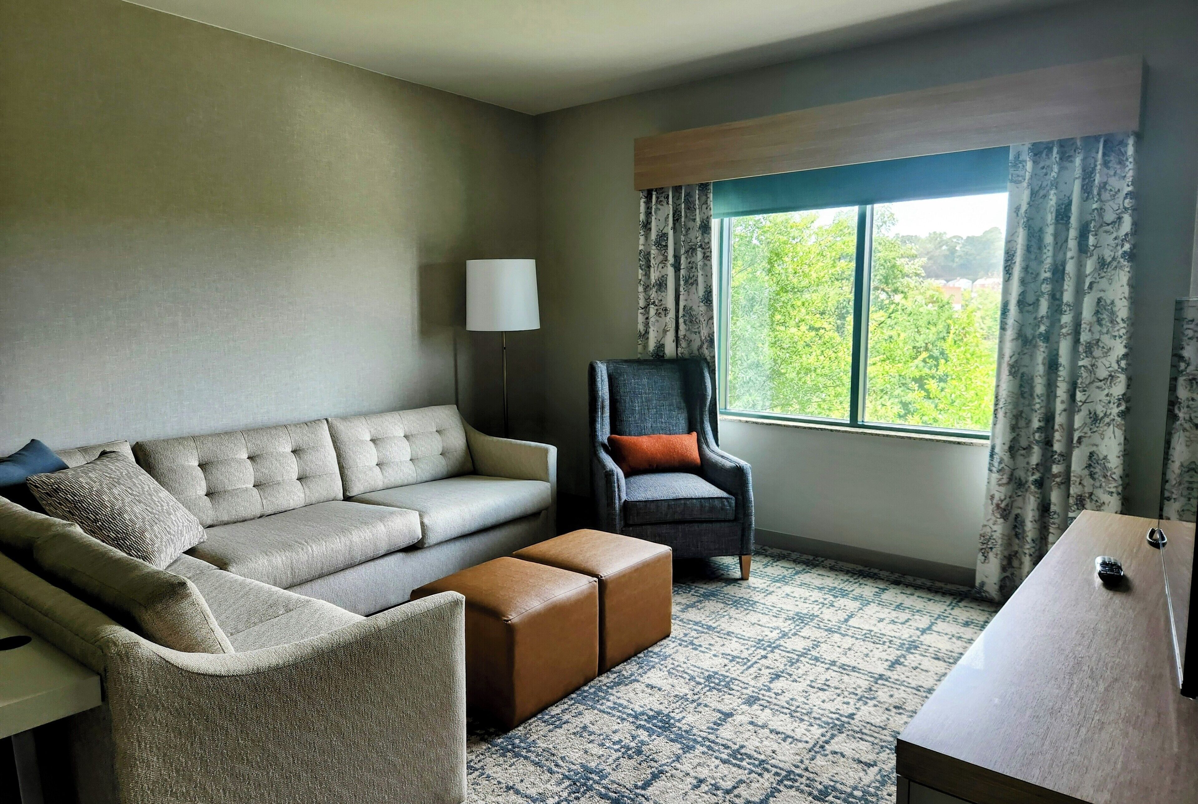 Un salon moderne avec un canapé, un fauteuil, un pouf et une vue sur la verdure par la fenêtre.