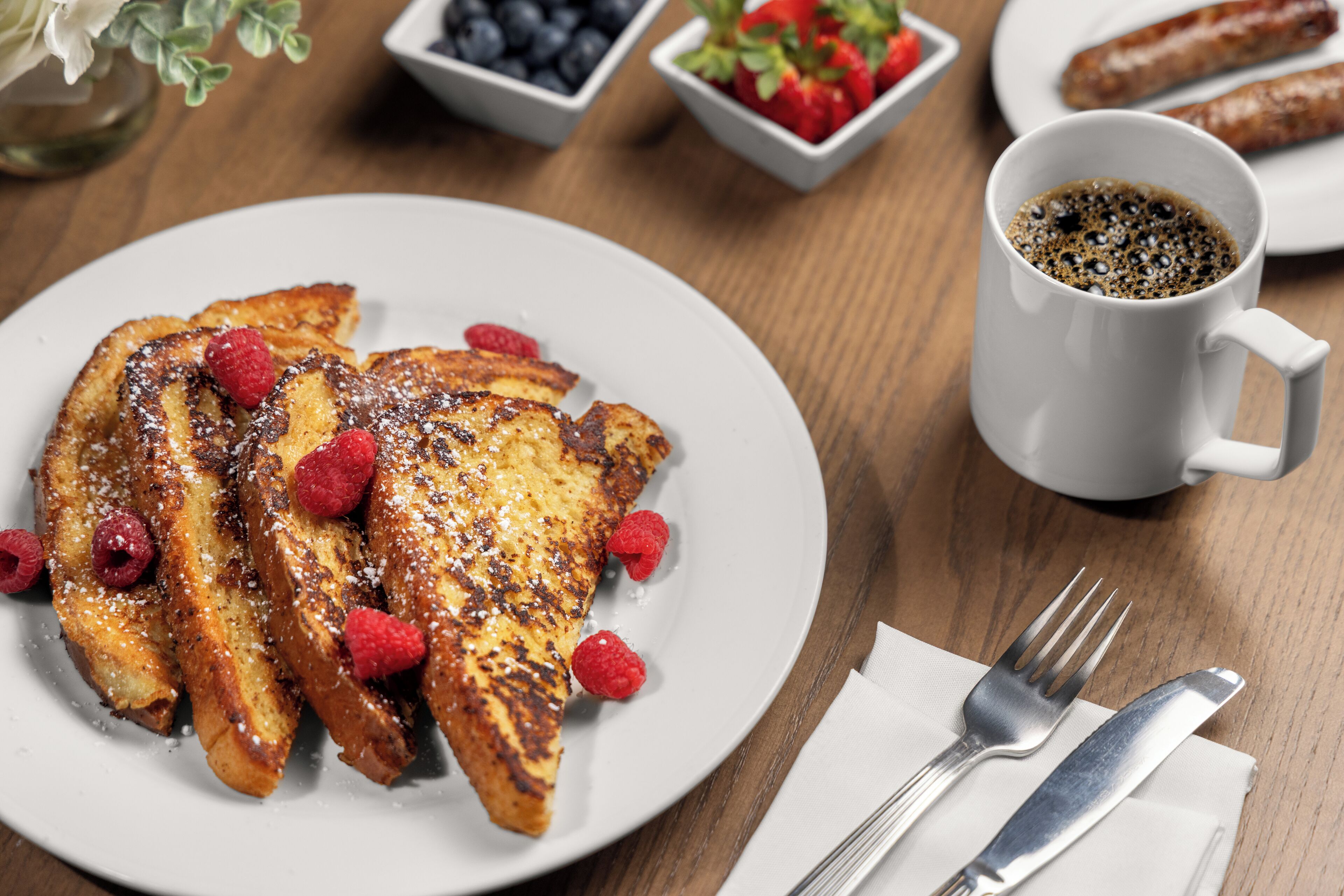 Ein Teller French Toast mit Himbeeren, eine Tasse Kaffee sowie eine Gabel und ein Messer auf einem Holztisch.