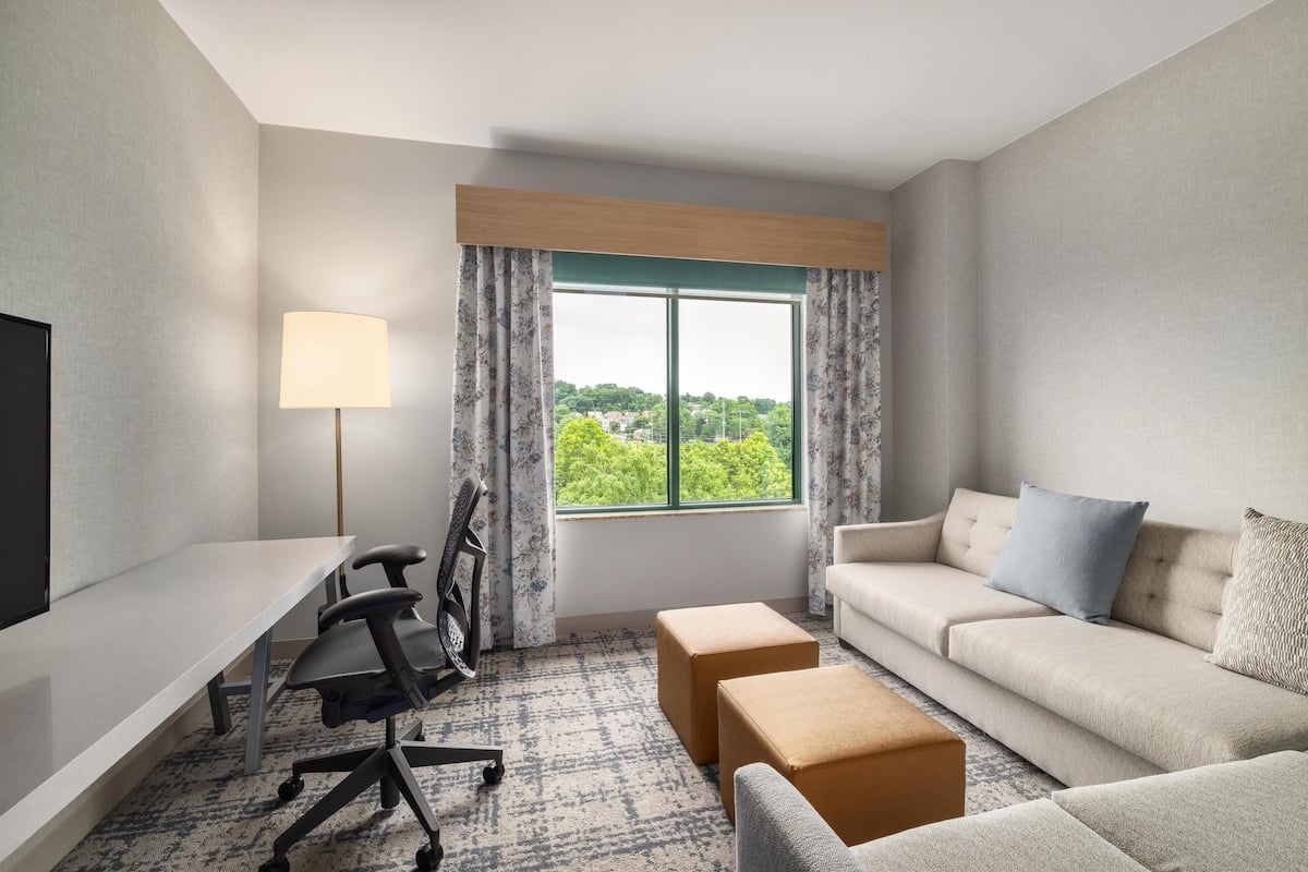 A modern hotel room with a desk, office chair, sofa, and ottomans.