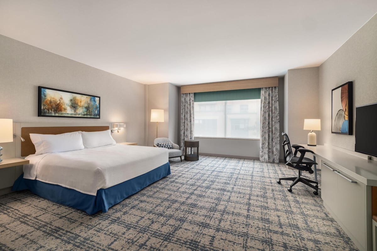 A hotel room with a large bed, a desk, a chair, a TV, and a window with curtains.