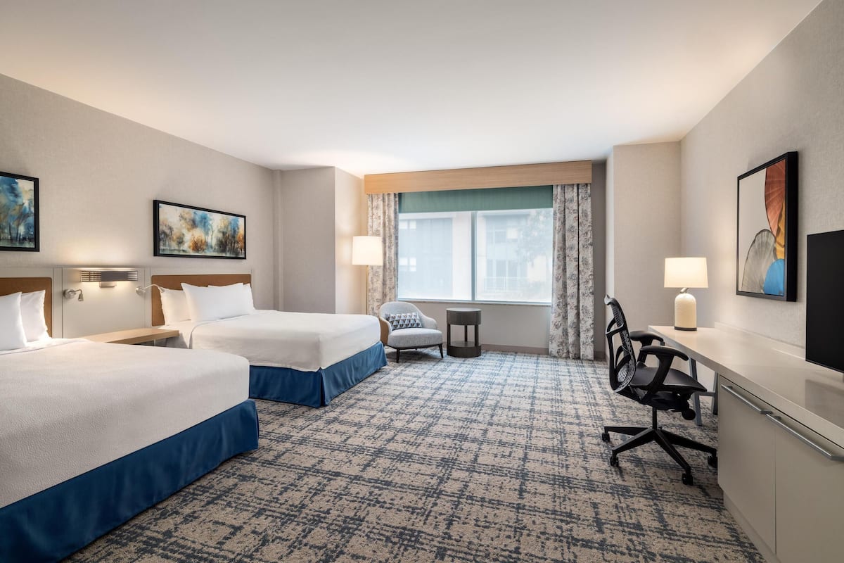 A hotel room with two beds, a desk, a chair, a lamp, and a window with curtains.