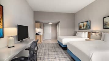 A hotel room with two beds, a desk with a chair, a TV, and a small kitchenette.