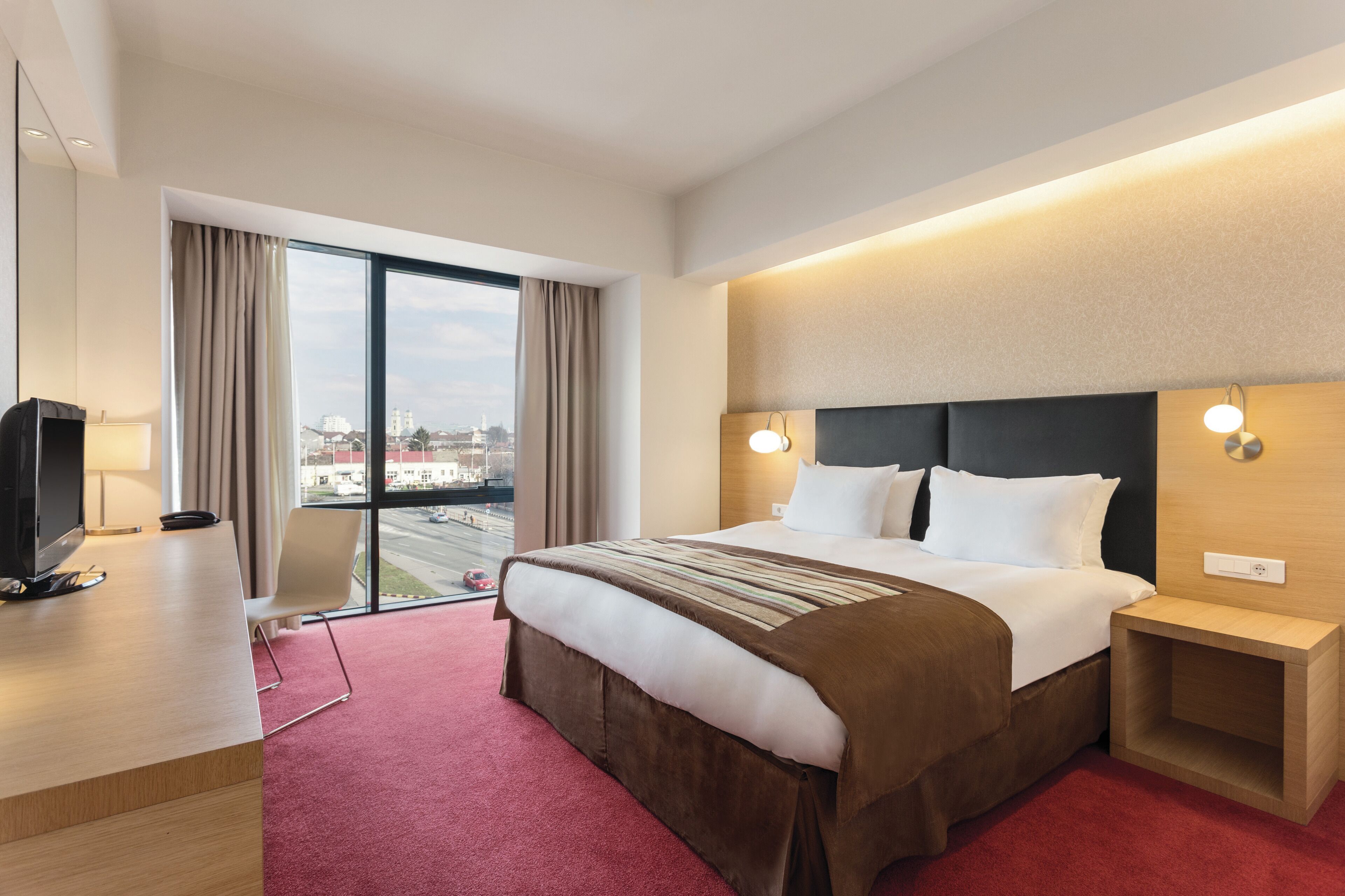A hotel room with a large bed, a desk, a TV, and a view of the city through the window.