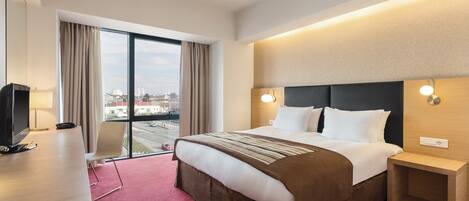 A hotel room with a large bed, a desk, a TV, and a view of the city through the window.