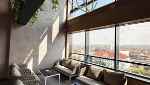 A modern lounge with wicker furniture, a glass coffee table, and a view of a cityscape through large windows. - Ramada by Wyndham Oradea (Oradea)
