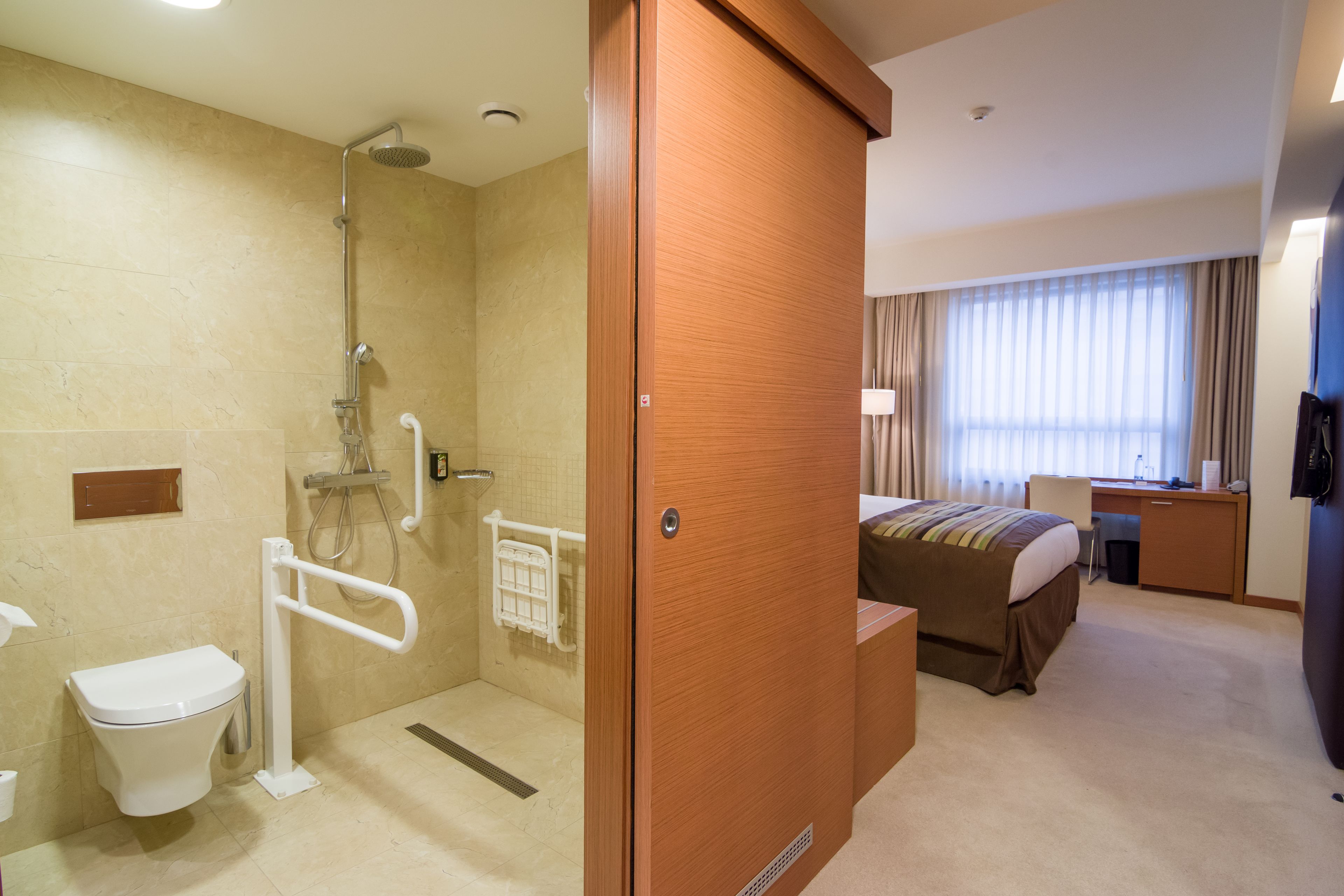 A hotel room with a shower, toilet, bed, desk, and a view of the outdoors.