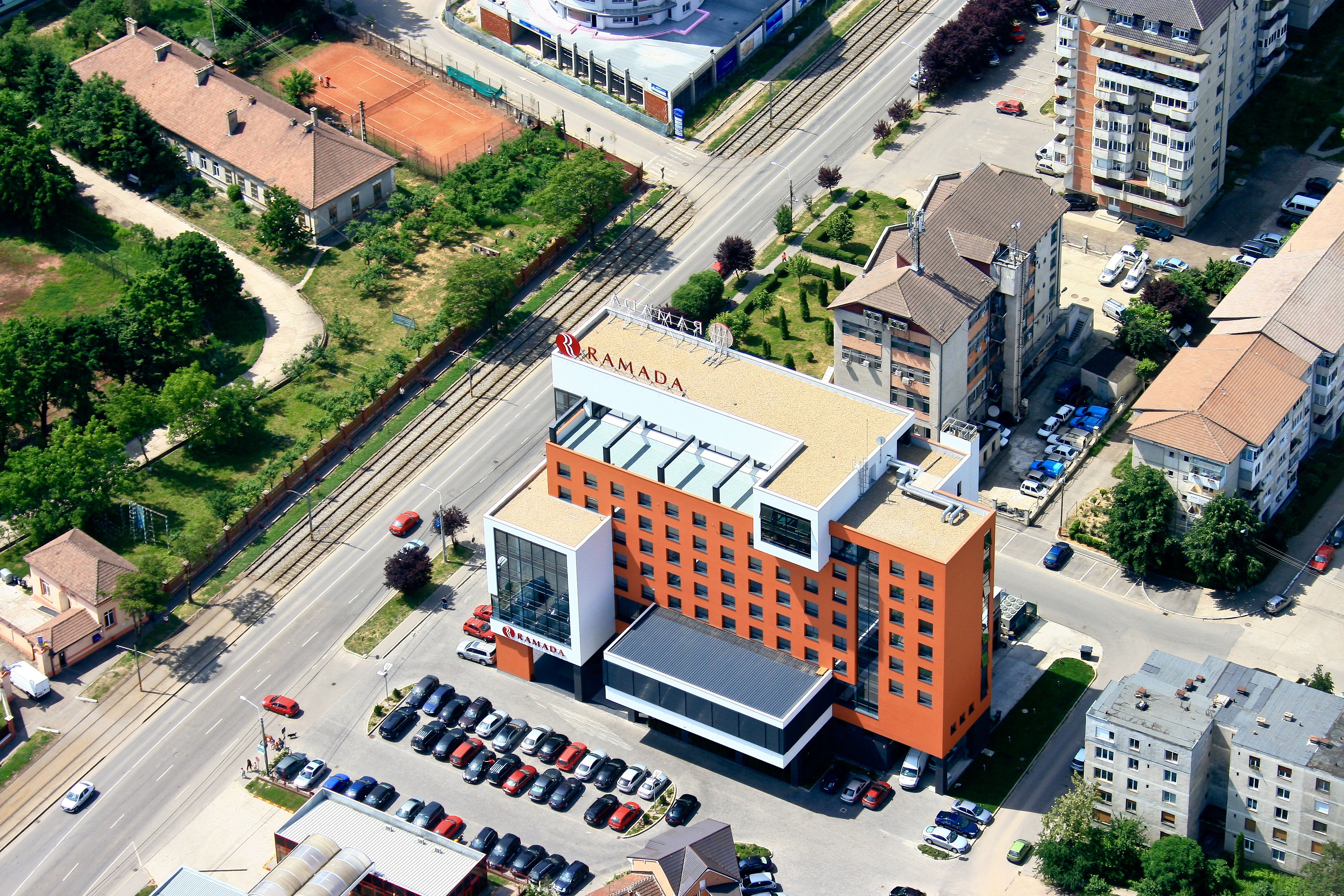 Un edificio de varios pisos con un llamativo letrero de 'RAMADA' en el techo, rodeado de estacionamiento y otros edificios.