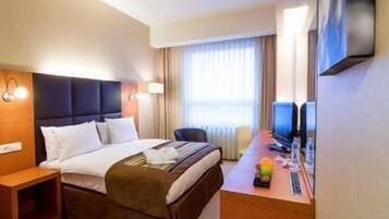 A hotel room with a bed, bedside table, desk with a computer, and a TV.