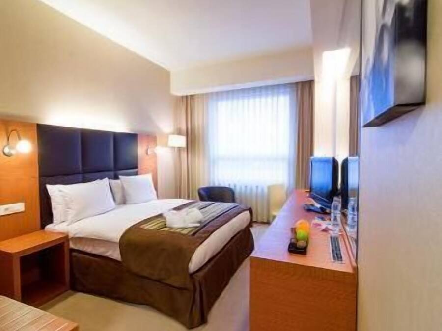 A hotel room with a bed, bedside table, desk with a computer, and a TV.