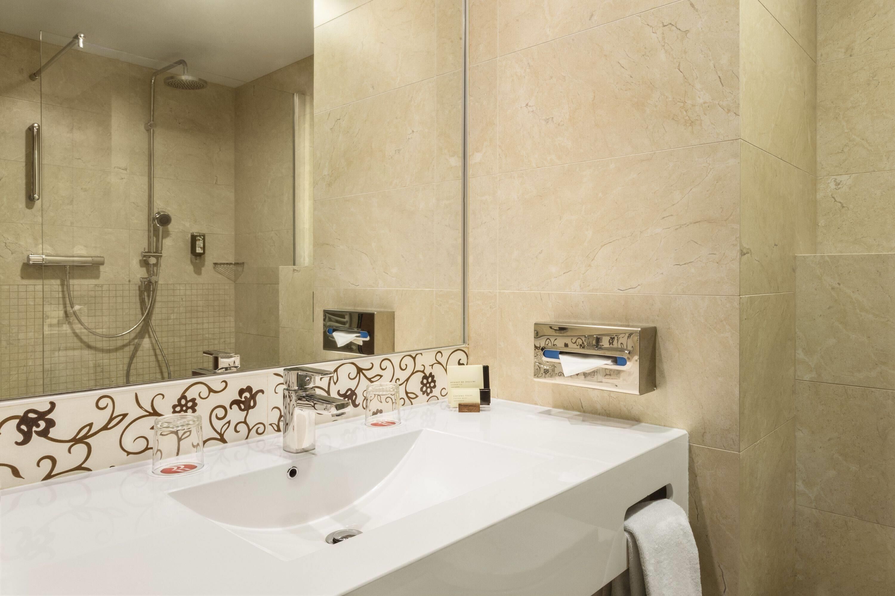A bathroom with a large mirror, a white sink, a handheld showerhead, and a towel rack.