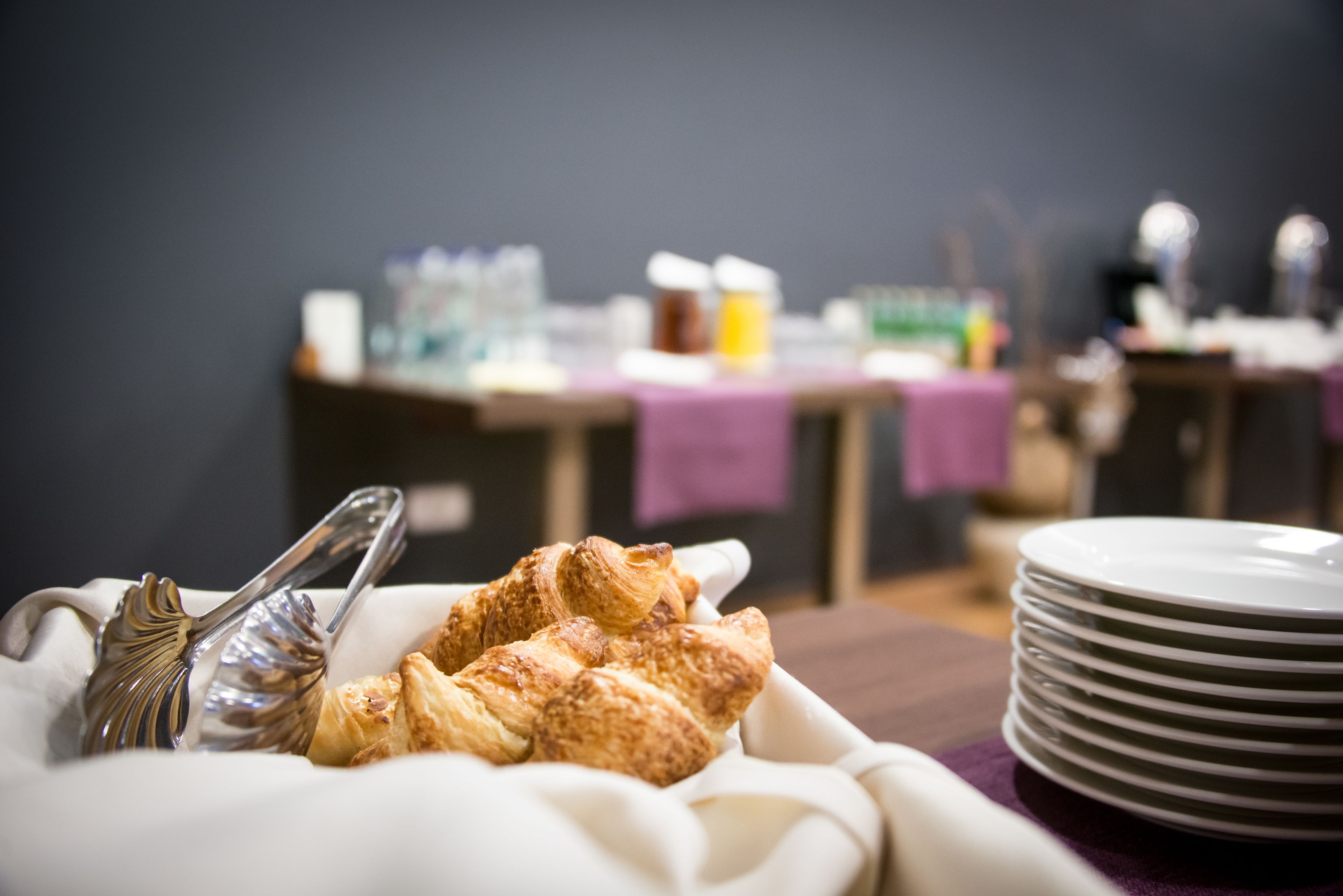 Une table mise pour le petit-déjeuner, avec des croissants et des couverts, et des assiettes empilées à proximité.