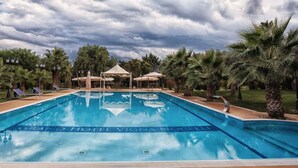 A swimming pool with lounge chairs and umbrellas, surrounded by palm trees and a cloudy sky. - Grand Hotel Vigna Nocelli (Lucera)