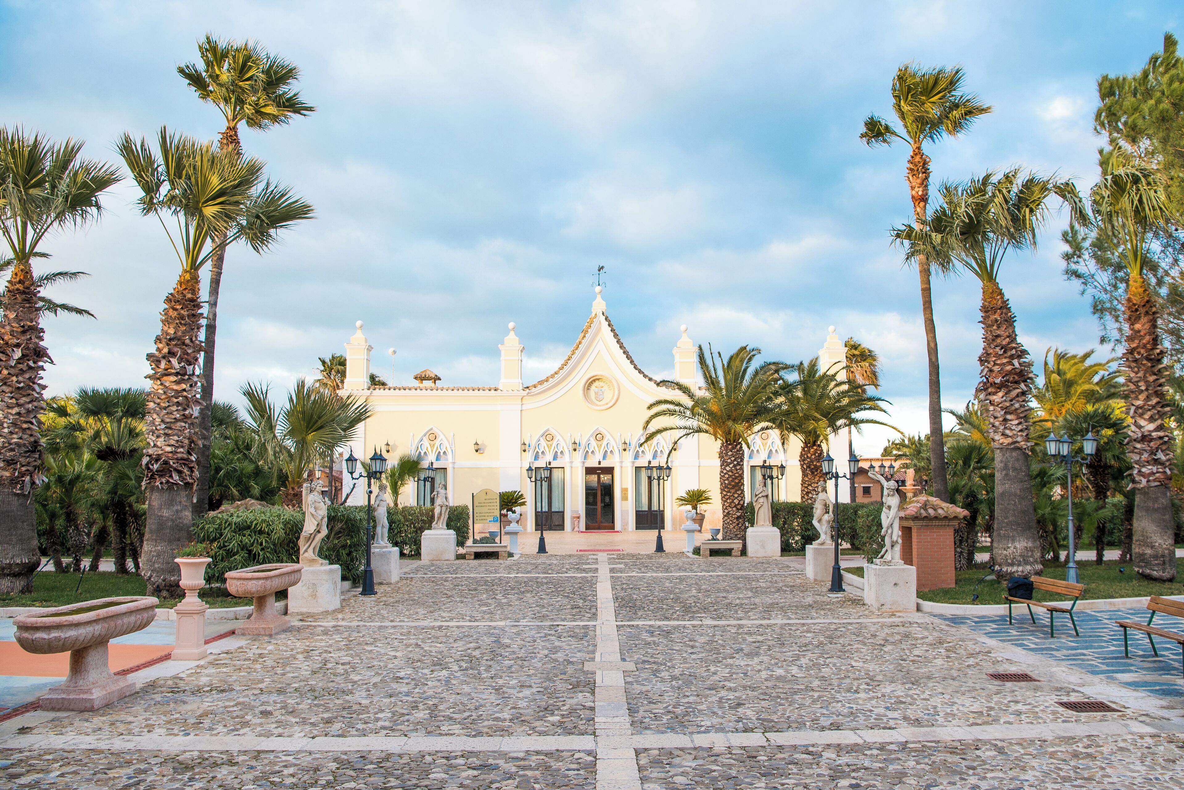 A courtyard with a building featuring a central dome and symmetrical design, surrounded by palm trees and stone planters.