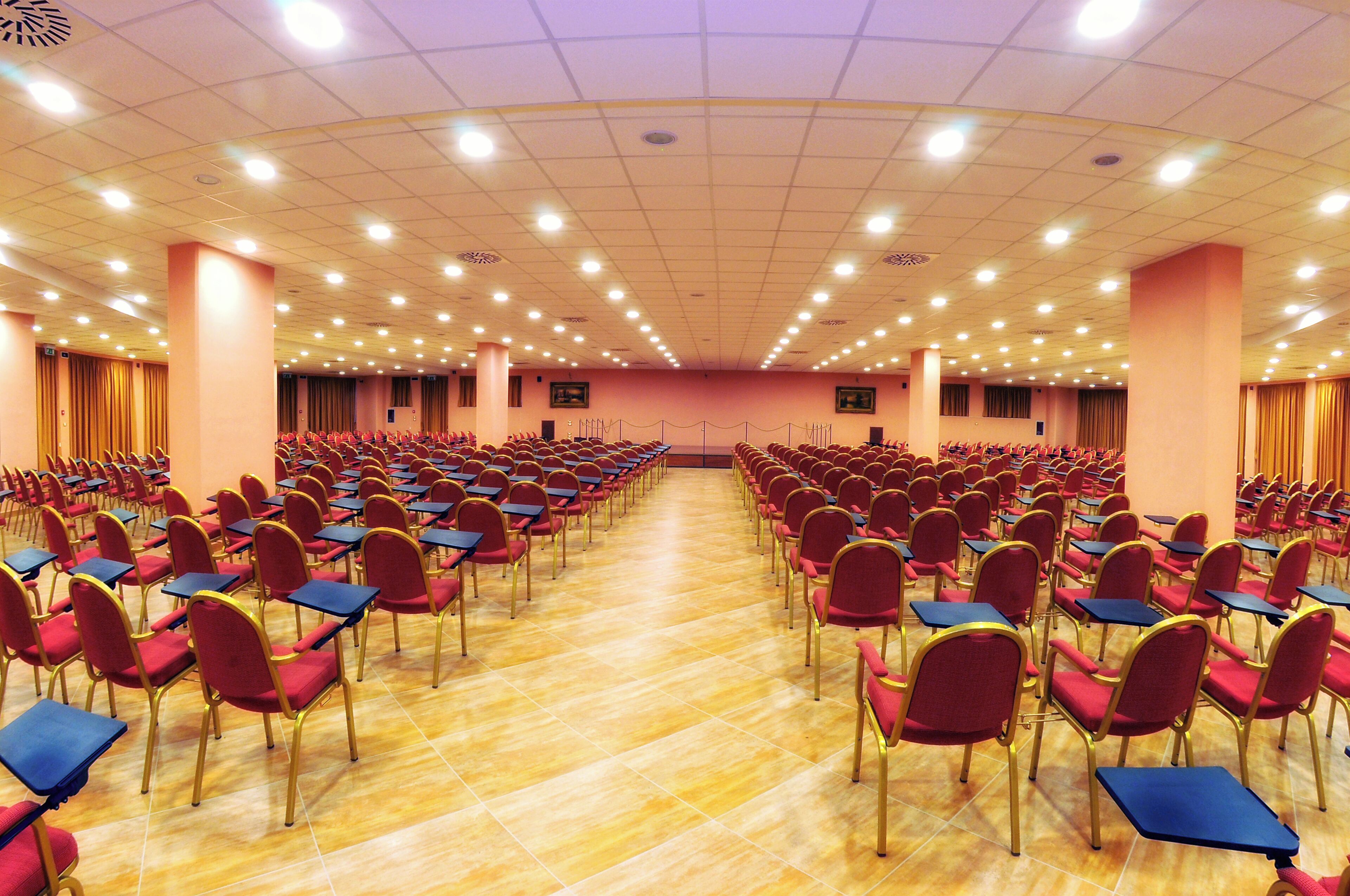 Un'ampia sala conferenze con file di sedie rosse e tavoli blu, disposti in modo simmetrico.