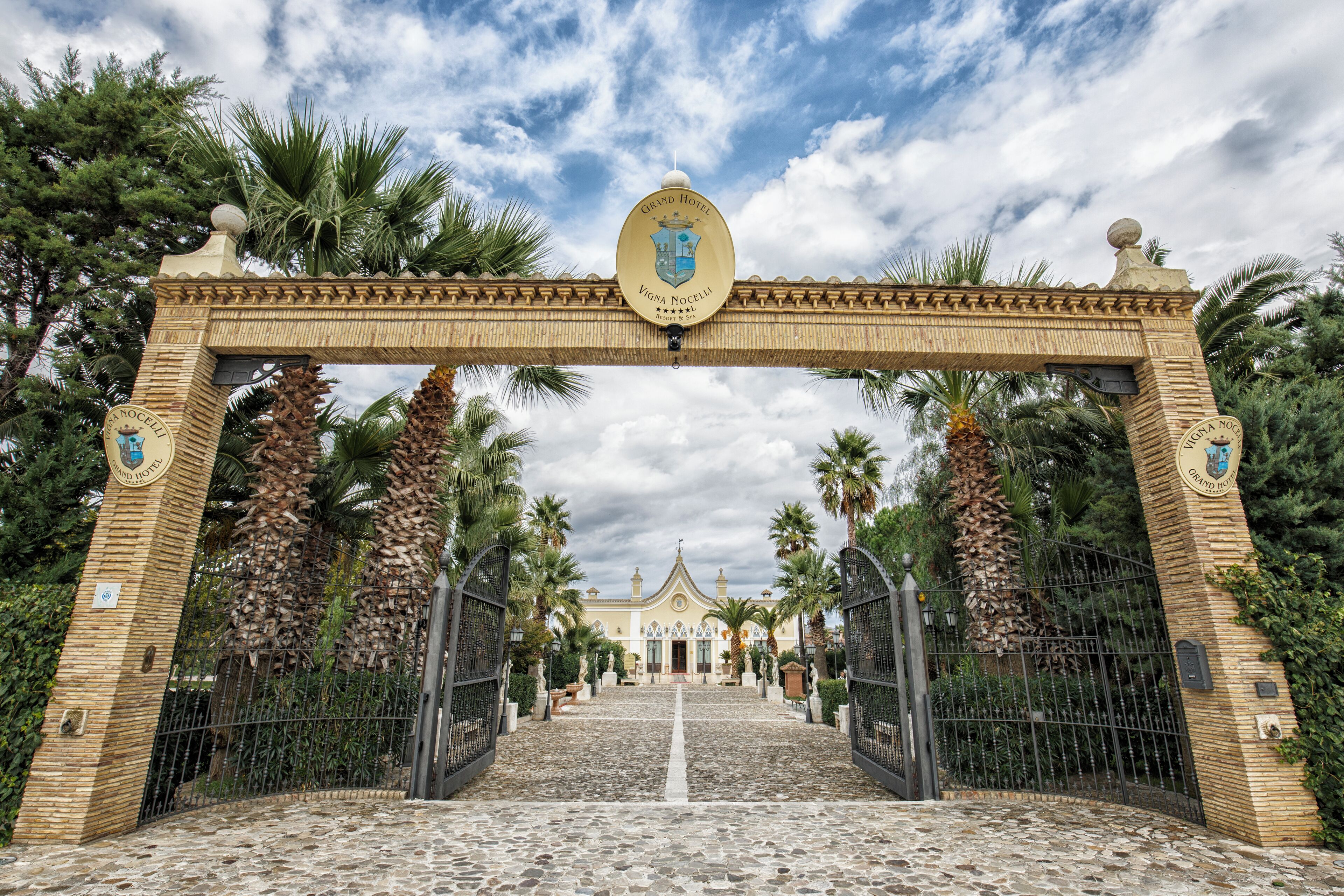 Una entrada imponente con un arco de piedra, palmeras y un edificio al fondo.