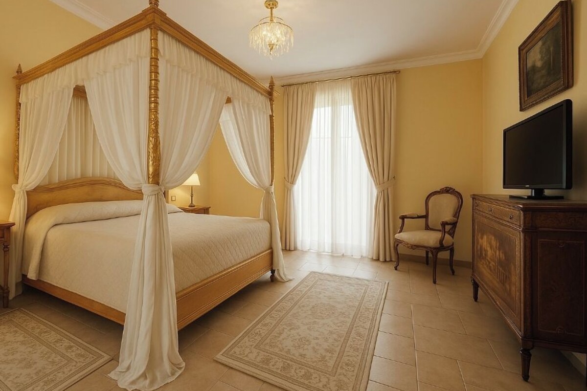 A four-poster bed with white drapes, a television on a wooden cabinet, and a chair in a room with beige walls and a chandelier.
