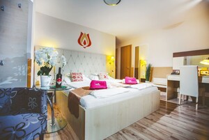Quarto de hotel com cama, escrivaninha, cadeira, pequena mesa com uma planta e um objeto decorativo na parede.