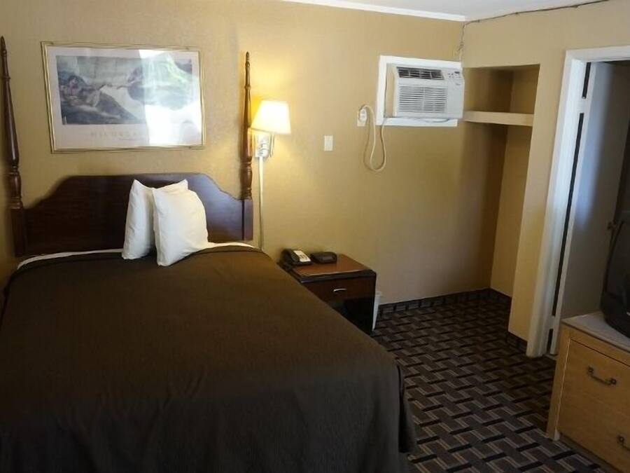 A hotel room with a bed, a nightstand, a lamp, an air conditioning unit, a television, and a wardrobe.