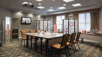 A conference room with a large table, chairs, a projector, and a large window with curtains.