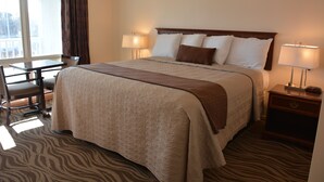 A hotel room with a large bed, two bedside lamps, a wooden nightstand, a desk with a chair, and a view of the outdoors through a window. - Liberty Inn (Old Saybrook)