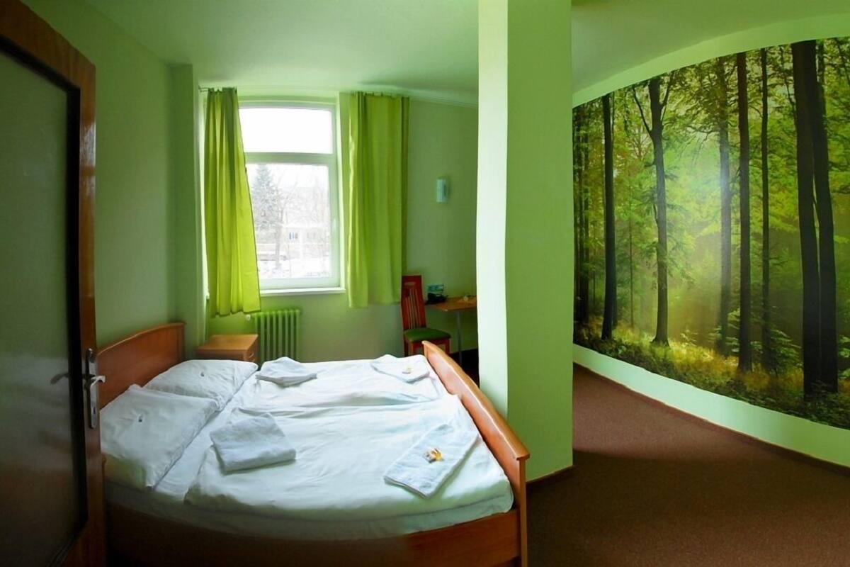 A bedroom with a bed, a window with curtains, a radiator, a desk, and a large forest mural on the wall.