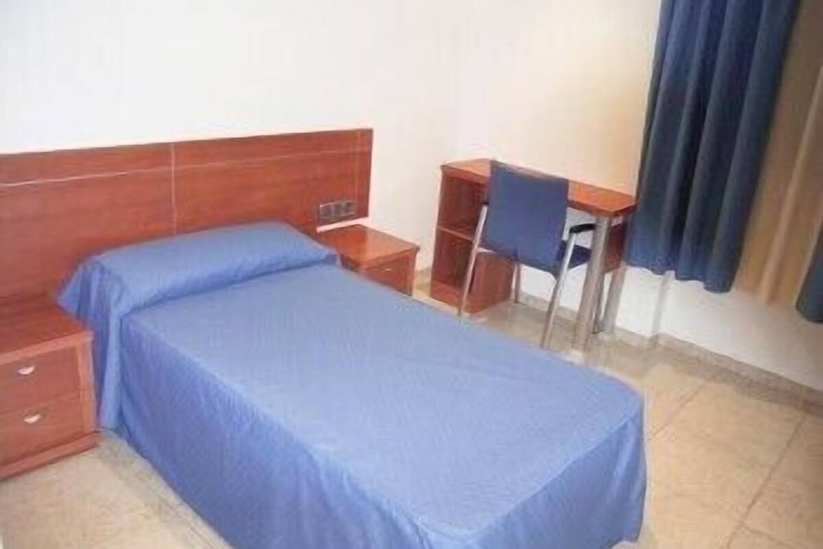 A single bed with a blue bedspread, a wooden nightstand, a desk with a chair, and a blue curtain.