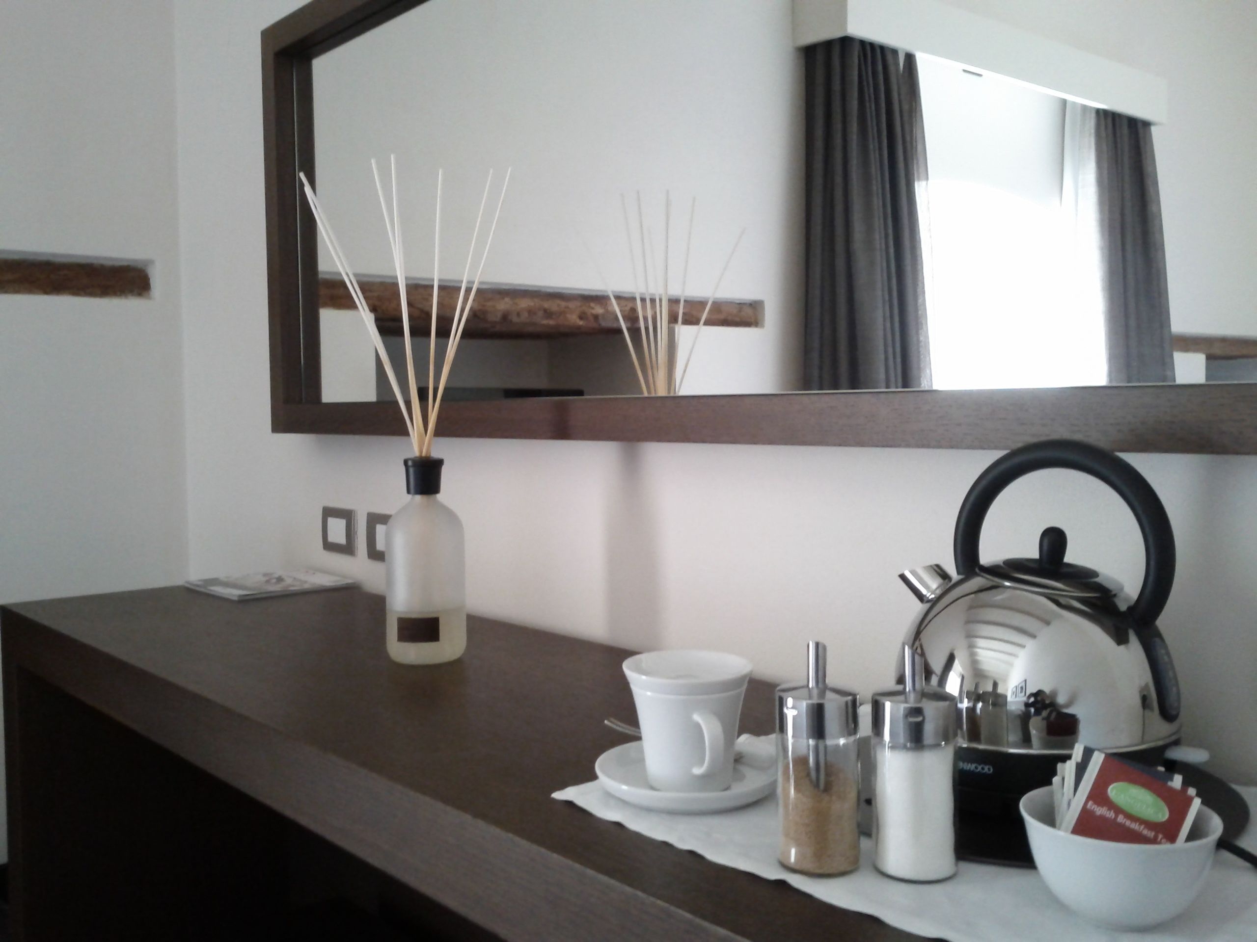 A hotel room with a dark wooden counter, a white vase with reed sticks, a white teapot, a white cup, a white saucer, a white bowl, and a mirror above the counter.