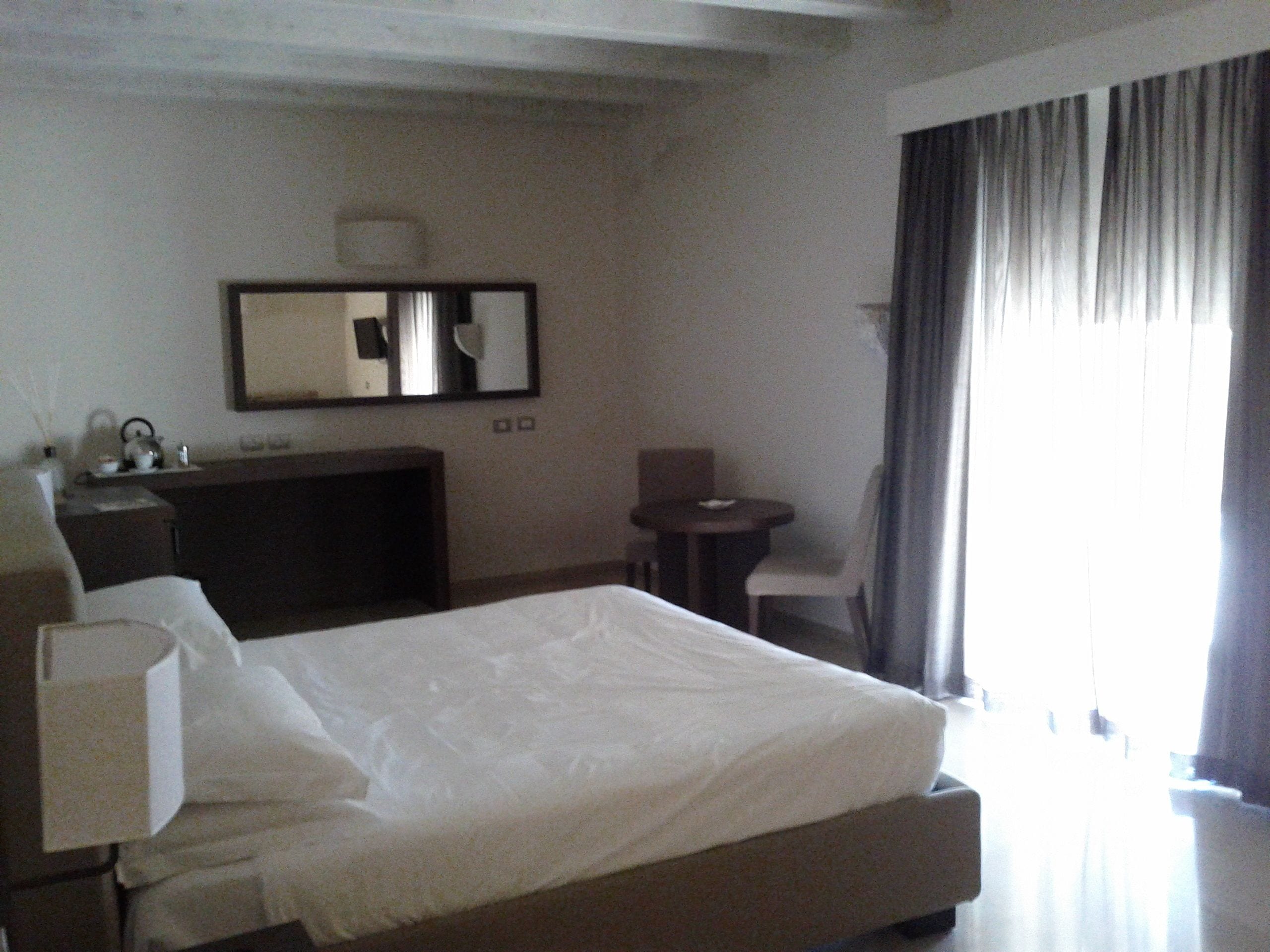 Une chambre d’hôtel avec un lit, un bureau, une chaise, une petite table et une grande fenêtre avec des rideaux.