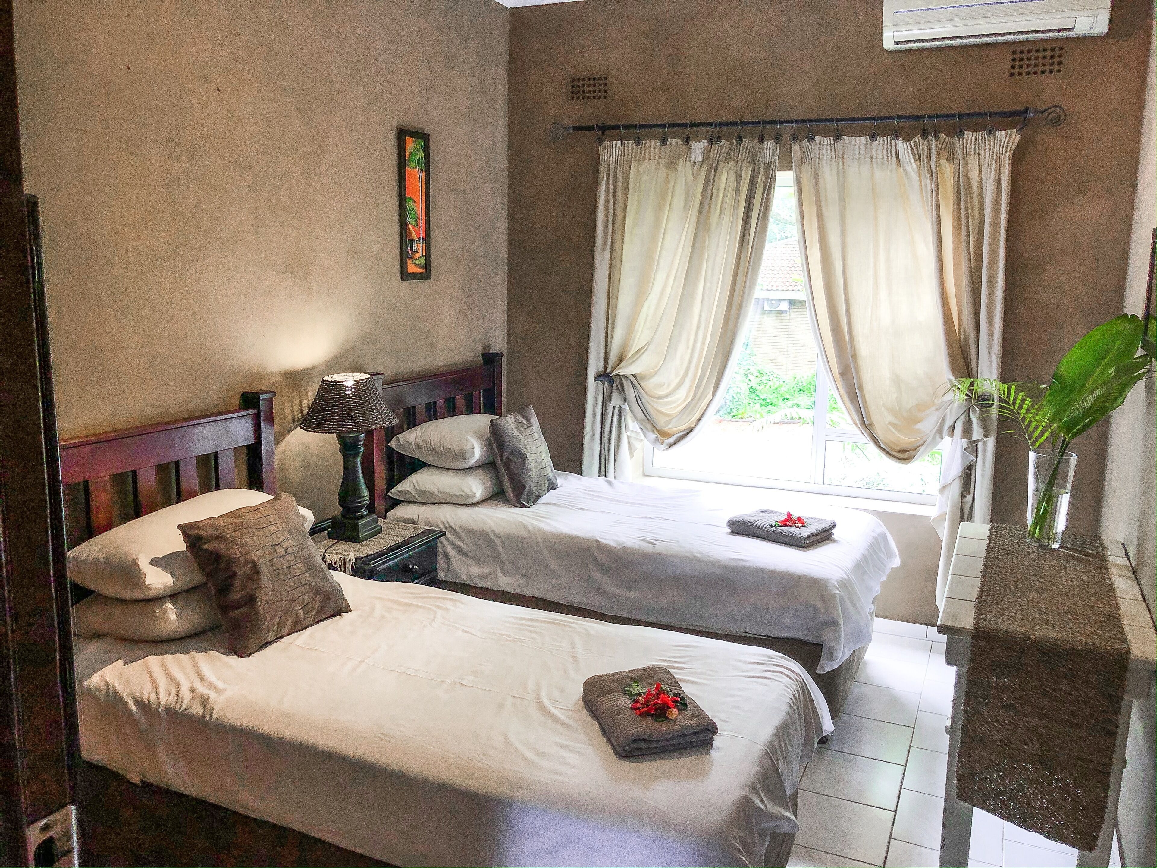 A room with two beds, a nightstand with a lamp, a window with curtains, and a plant on a table.