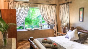 A bedroom with a large bed, a window with curtains, a bedside table with a lamp, and a potted plant. - Ingwenya Lodge (St. Lucia)
