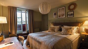 A bedroom with a large bed, a desk, and a chair. There is a window with curtains, a wall-mounted lamp, and framed pictures.