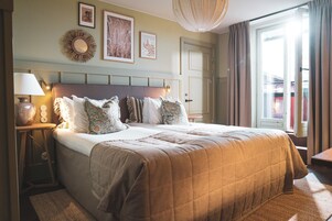 Quarto de hotel com uma cama grande, mesa de cabeceira com abajur, relógio na parede e janela com cortinas.