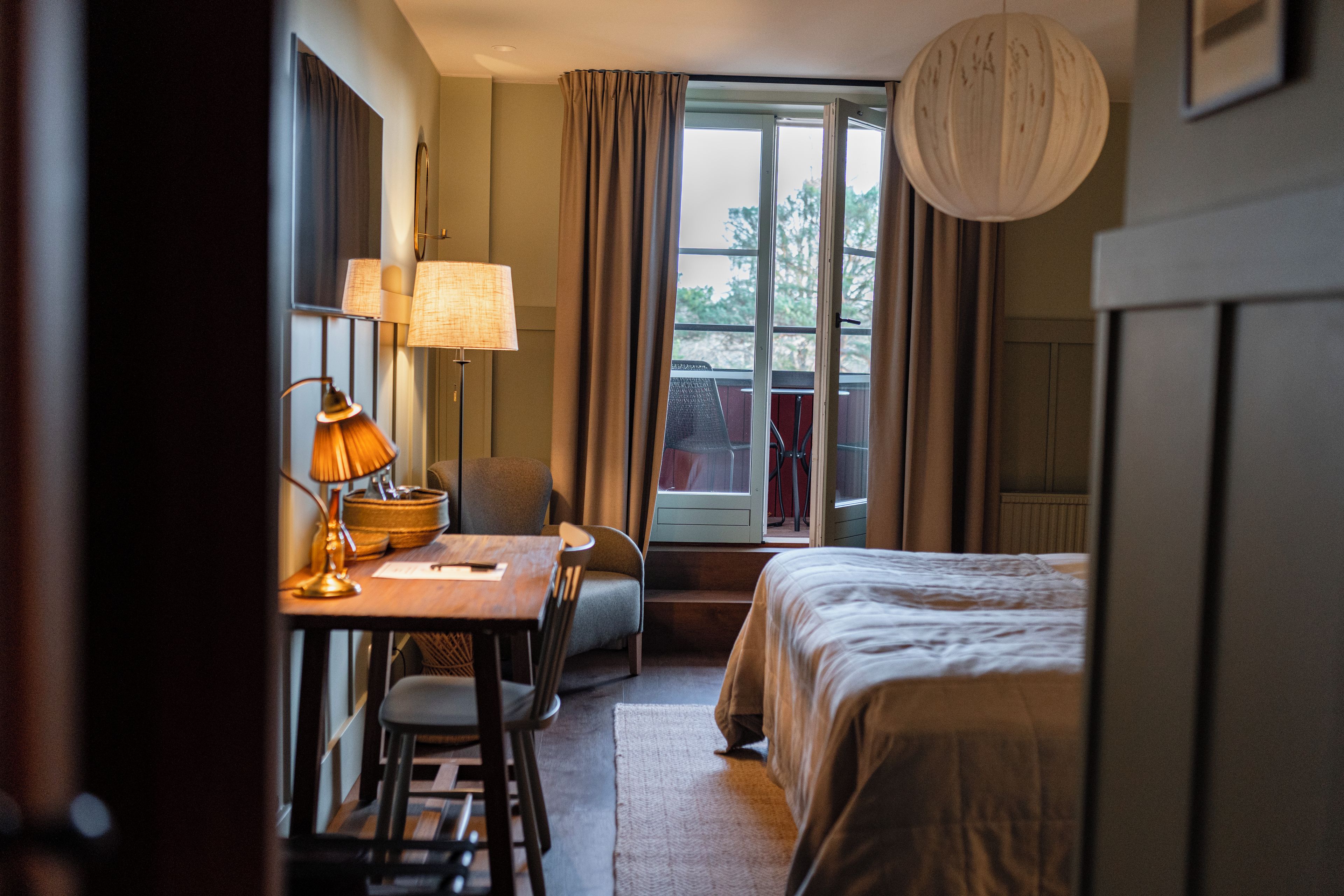 A hotel room with a bed, a desk with a lamp, a chair, and a view of the outdoors through a window.