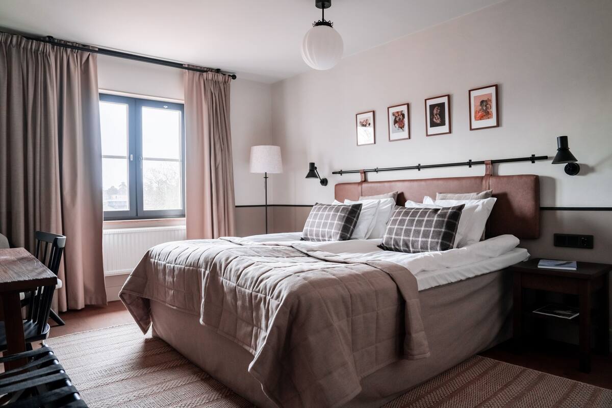 Un dormitorio ordenado con una cama grande, una mesita de noche, una lámpara y una ventana con cortinas.
