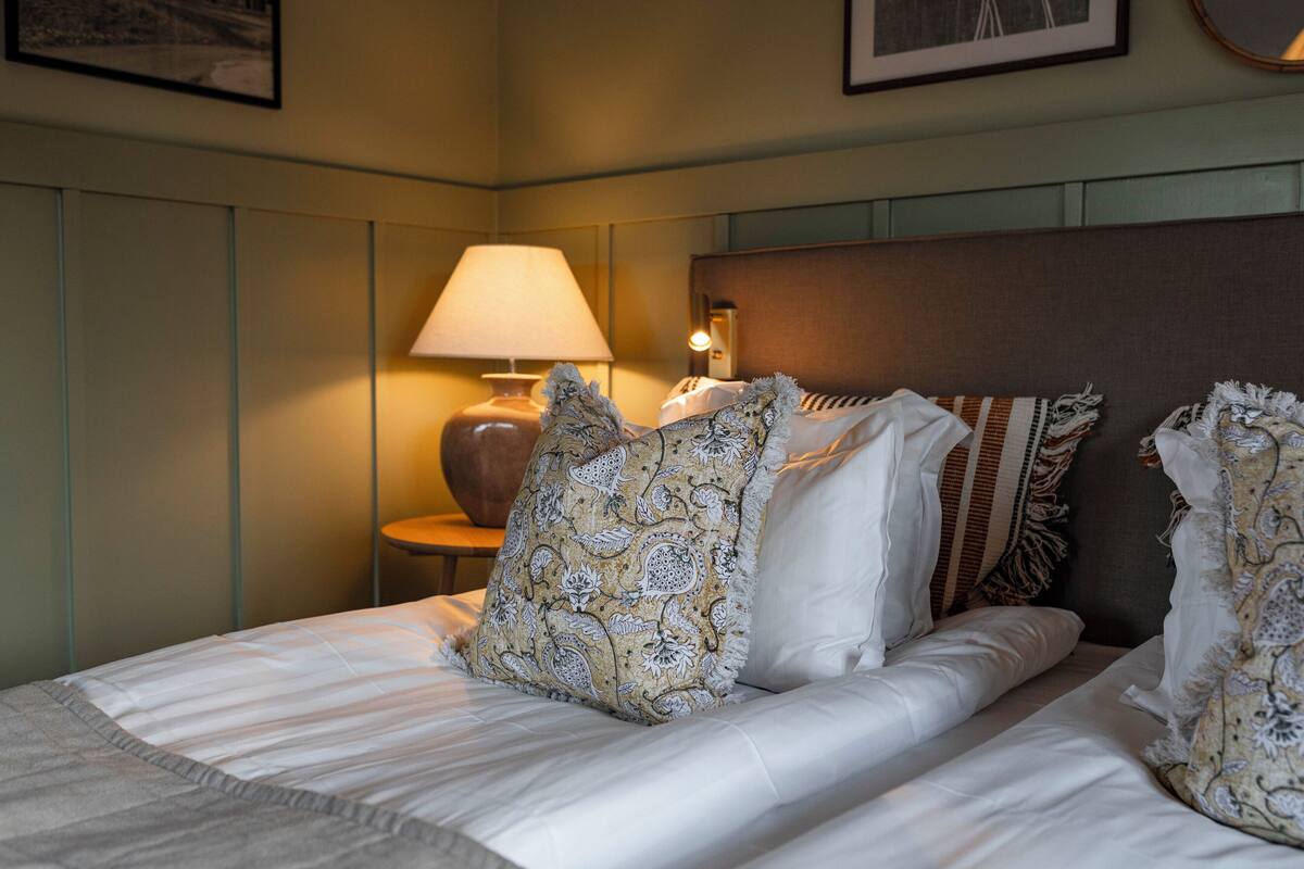 Una cama bien tendida con almohadones estampados, una lámpara de noche y un cuadro en la pared.