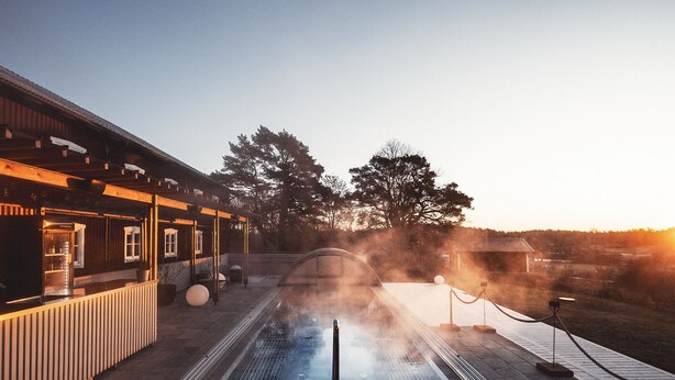 A spa pool with a wooden deck, a covered seating area, and a view of trees and a sunset. - Fågelbro Säteri (Fågelbrolandet)