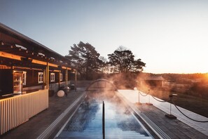 A spa pool with a wooden deck, a covered seating area, and a view of trees and a sunset. - Fågelbro Säteri (Fågelbrolandet)