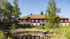 A large, two-story building with a red facade and a tiled roof, featuring a bell tower and a covered outdoor seating area. - Fågelbro Säteri (Fågelbrolandet)